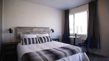 Une chambre d’hôtel avec un grand lit, une tête de lit, deux tables de chevet, une petite table ronde, deux chaises et une grande fenêtre avec des rideaux.