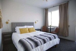 A hotel room with a bed, a small table, two chairs, a window with curtains, and wall-mounted lights.