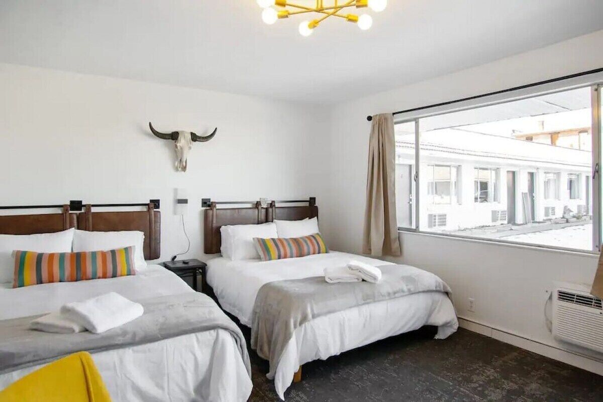 A hotel room with two beds, a large window with a view of buildings, a cow skull decoration on the wall, and a modern chandelier.