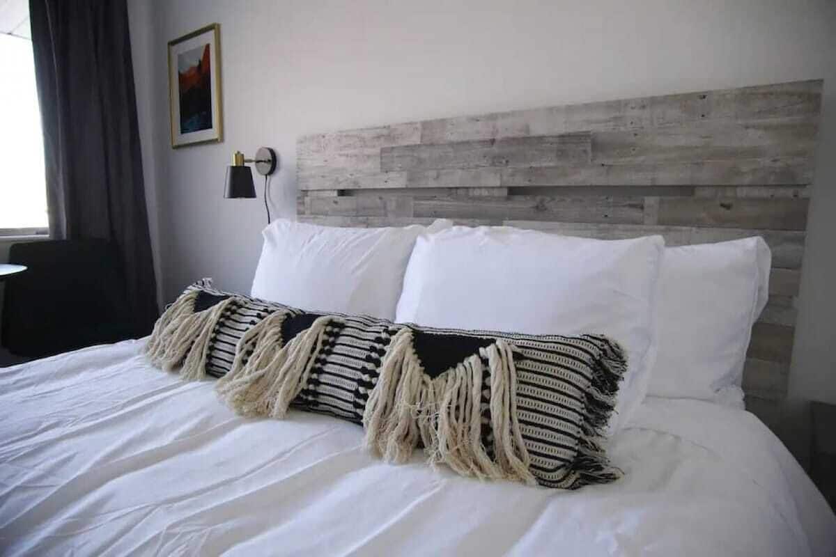A bed with white linens and a striped throw pillow, a wooden headboard, and a wall-mounted lamp.