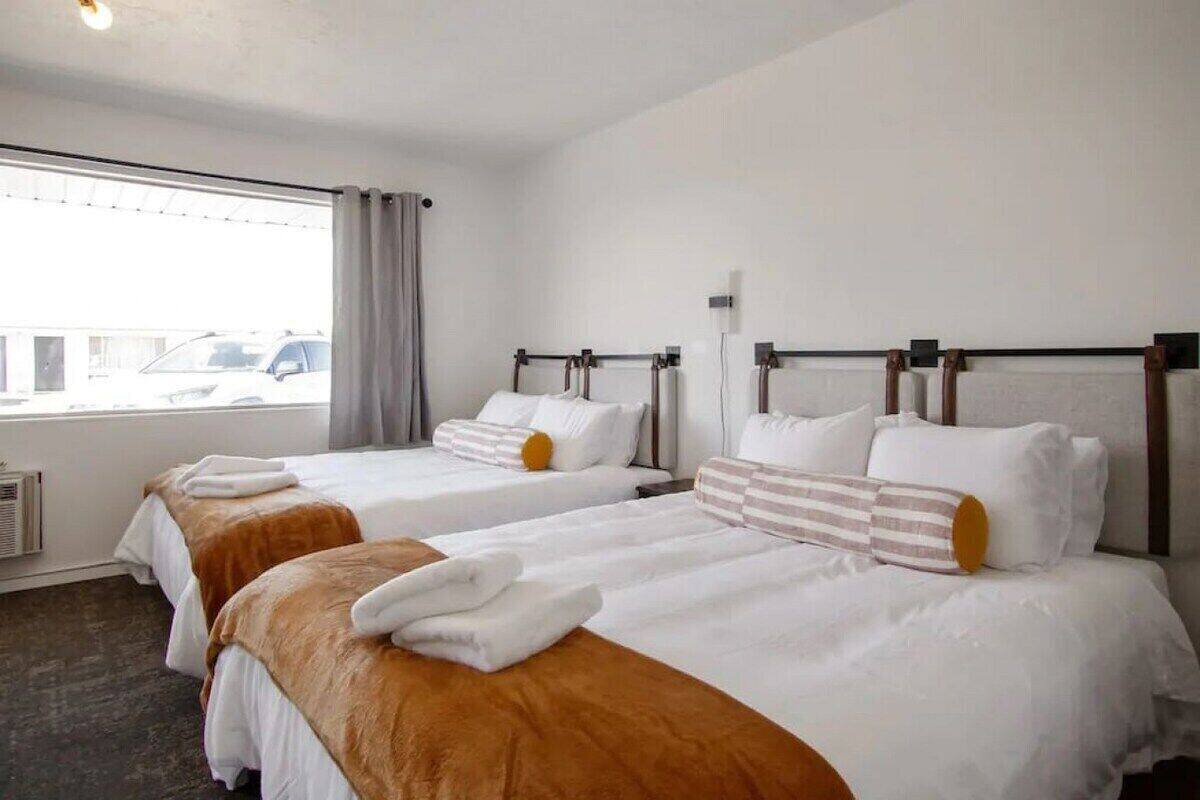 A hotel room with two beds, each with white bedding and brown headboards. There is a window with a view of parked cars outside.