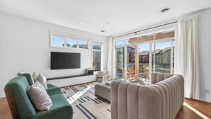A modern living room with a flat-screen TV, green and beige sofas, a patterned rug, and a large sliding glass door leading to a balcony. - Lakefront Pool and Lake View 2 Bdrm 4205 (West Kelowna)