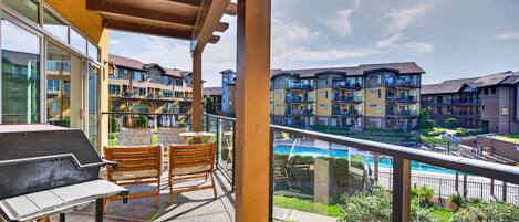 A balcony with a grill, chairs, and a view of multi-story buildings and a swimming pool.