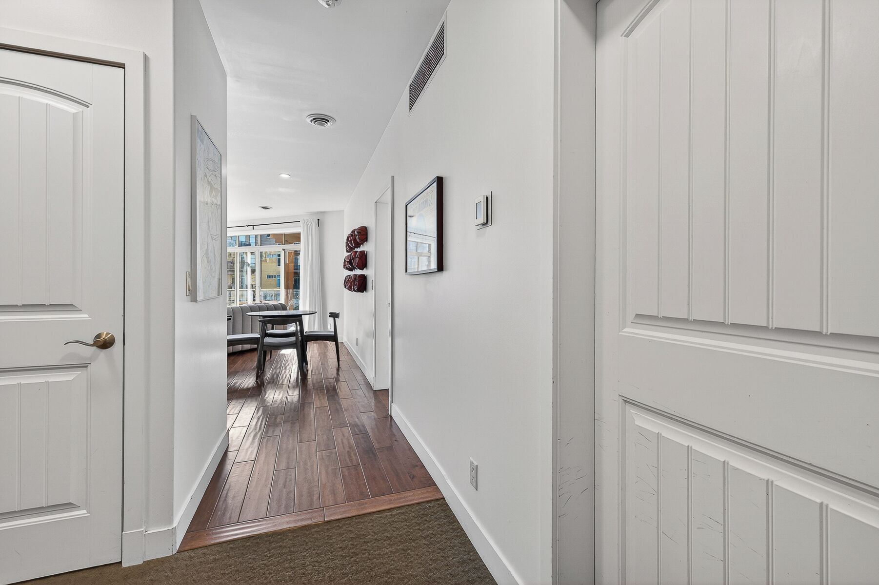 A hallway with white paneling, a dining area with a table and chairs, and a view of the outside through a window.