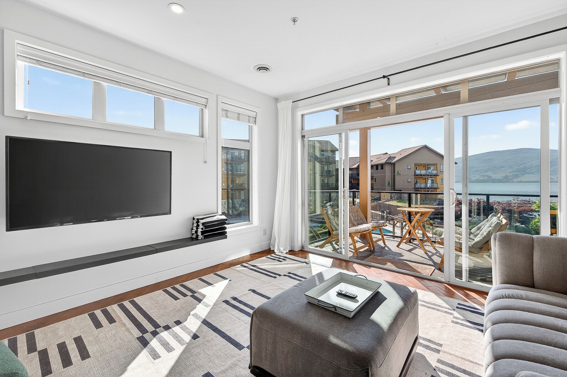 A spacious living room with a large flat-screen TV, a comfortable sofa, a small table, and a balcony with a view of buildings and a body of water.