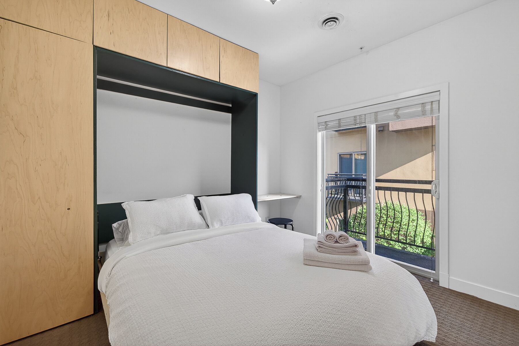 A bedroom with a bed, a wooden wardrobe, a small table, and a balcony with a railing.