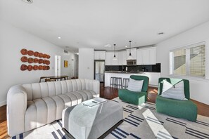Un salon moderne avec un canapé, deux fauteuils verts, une table à manger et une cuisine en arrière-plan.