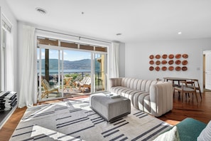A modern living room with a sofa, ottoman, and a dining table with chairs. - Lakefront Pool and Lake View 2 Bdrm 4205 (West Kelowna)