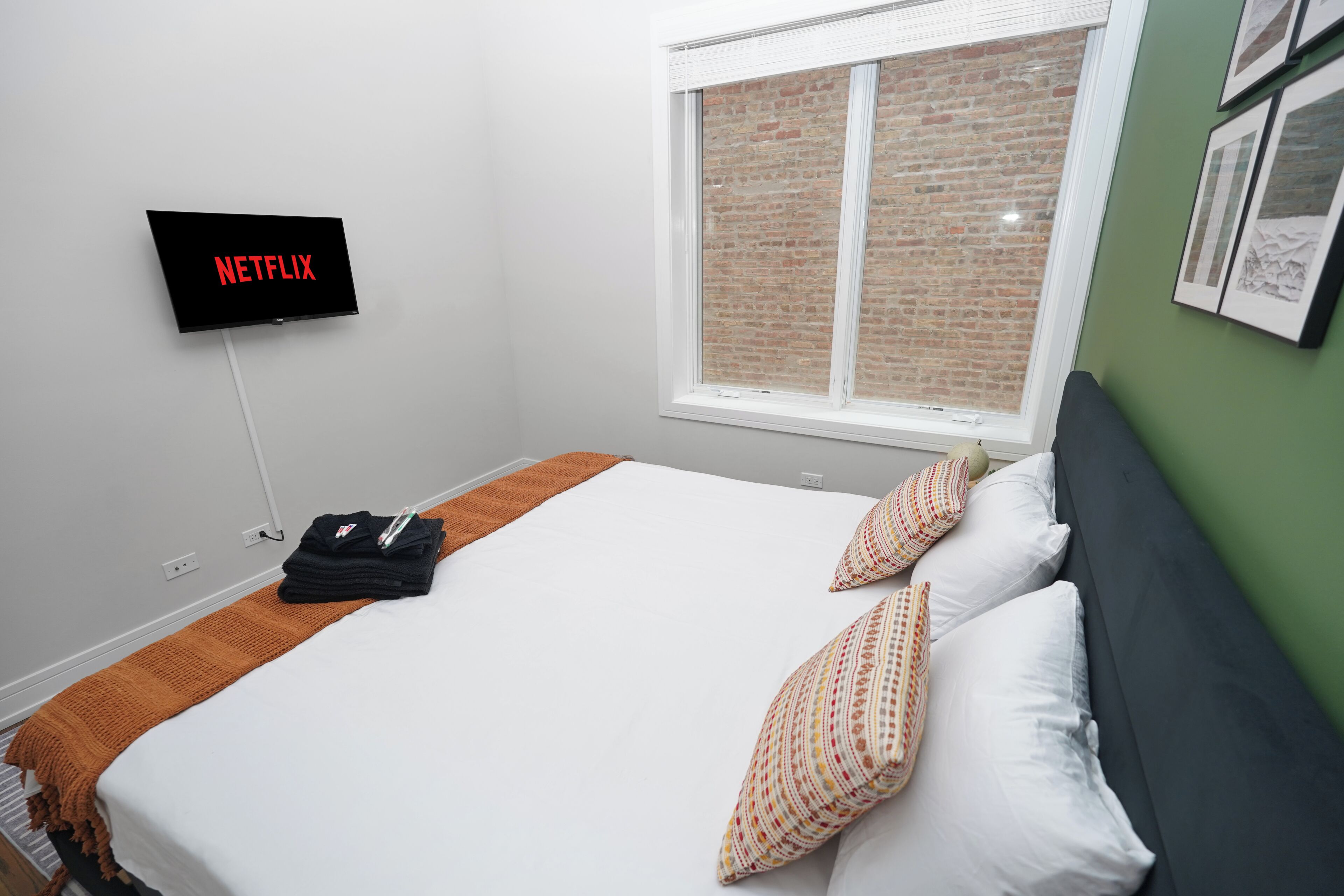 A hotel room with a bed, pillows, a blanket, a TV displaying 'NETFLIX,' a window with a brick wall view, and a green wall with framed pictures.