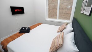 Een hotelkamer met een bed, kussens, een deken, een tv waarop 'NETFLIX' staat, een raam met uitzicht op een bakstenen muur, en een groene wand met ingelijste foto’s.