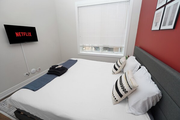 A neatly made bed with a white comforter and pillows, a blue blanket, a wall-mounted TV displaying 'NETFLIX,' and a window with blinds. - Spacious 4BR West Loop Retreat w/Optional Parking (Chicago)