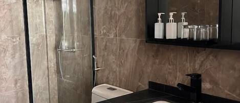 Un baño con ducha de vidrio, inodoro blanco, tocador de mármol negro y espejo sobre el lavabo.