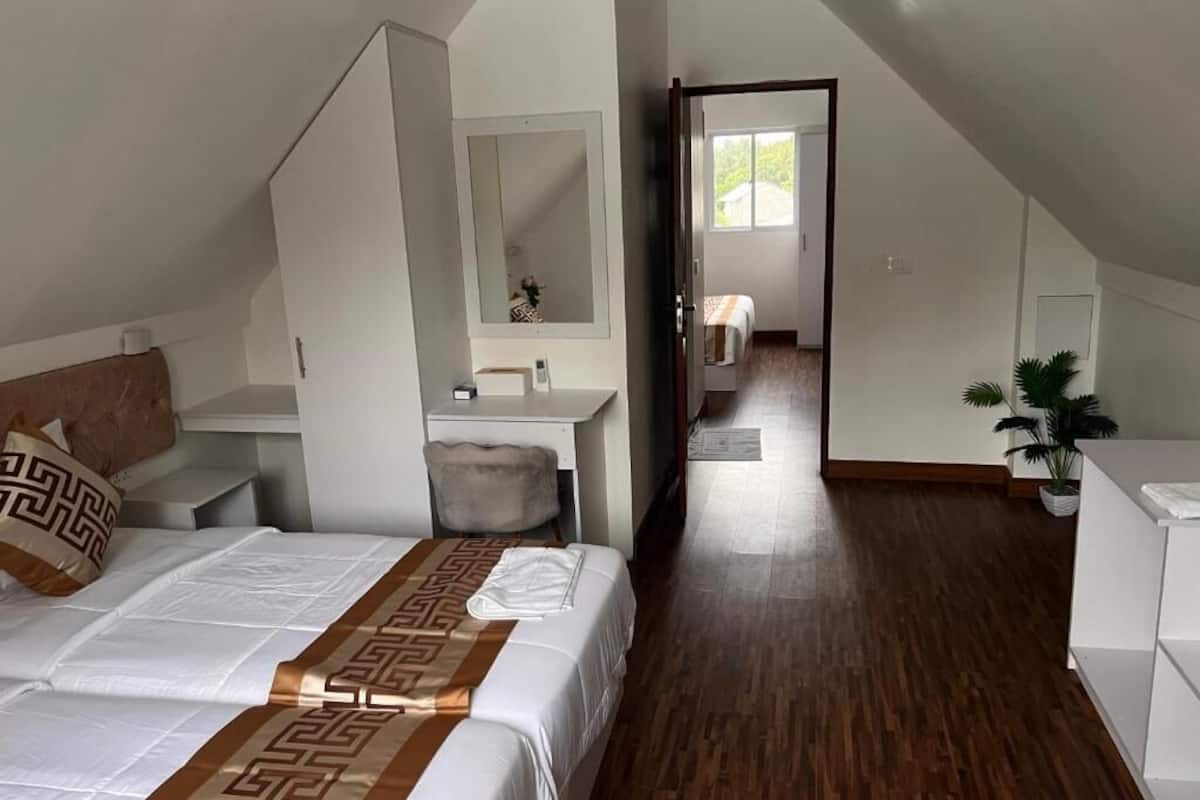 Une chambre avec un lit, une table de chevet et un miroir. Un couloir mène à une salle de bain équipée d’un lavabo et agrémentée d’une plante en pot.