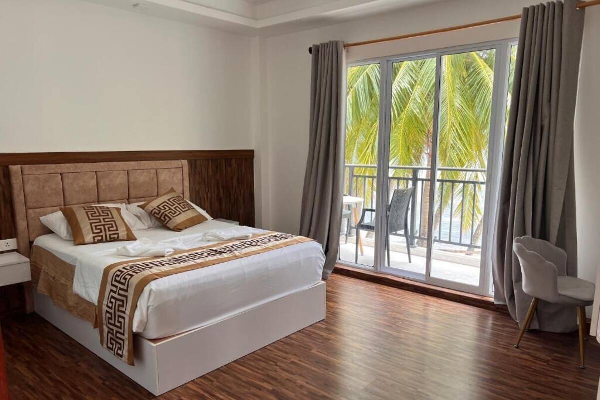 Une chambre d’hôtel avec un grand lit, un balcon donnant sur des palmiers et un fauteuil.