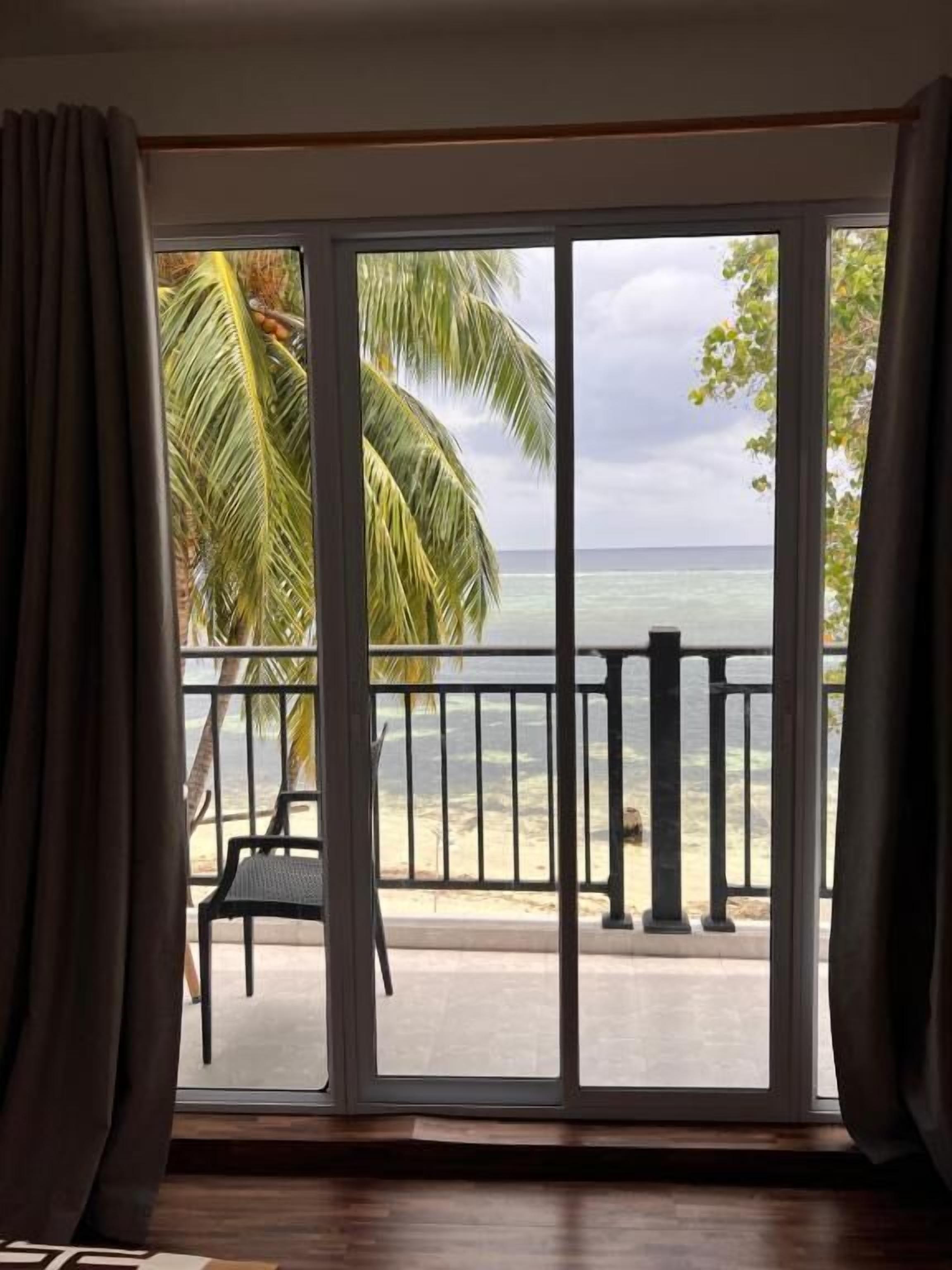 A room with a large sliding glass door leading to a balcony with a view of the ocean and palm trees.