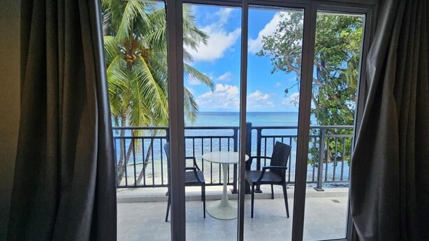 A room with a large sliding glass door leading to a balcony with a view of the ocean and palm trees. - Alma Maldiva beach hotel (Dhidhdhoo)