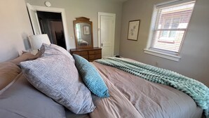 A bedroom with a bed, pillows, a dresser, a mirror, a window with blinds, and a door. - Stone's Throw Bungalow - Cozy and Convenient in Chambersburg, PA (Chambersburg)