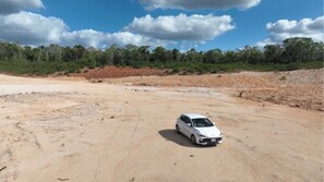 Une voiture blanche est garée sur un chemin de terre, avec une végétation clairsemée et des arbres en arrière-plan.