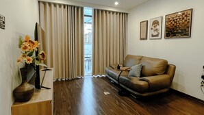 A living room with a leather sofa, wooden flooring, a vase with flowers, and a view through the curtains. - 2BR 2BTR Penthouse w/ Kitchen & BBQ Terrace in Long Bien (Hà Nội)