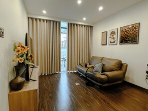A living room with a leather sofa, wooden flooring, a vase with flowers, and a view through the curtains. - 2BR 2BTR Penthouse w/ Kitchen & BBQ Terrace in Long Bien (Hà Nội)