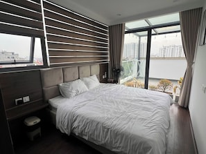 Ein modernes Schlafzimmer mit einem großen Bett, einem Nachttisch, einem Ventilator und Blick auf eine Stadtlandschaft durch ein großes Fenster.