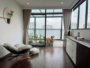 A modern living room with a rocking chair, a dining area with a coffee maker, and a view of the cityscape through large windows. - Penthouse tầng 7 2BR và Ban công lớn cho tiệc BBQ (Hà Nội)