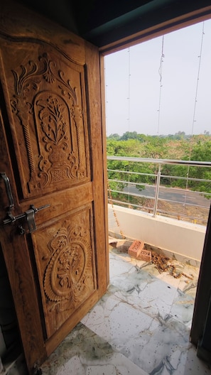 An open wooden door with intricate carvings, leading to a balcony with a view of a road and greenery. - Sandhya Guest House (Ekangardih)