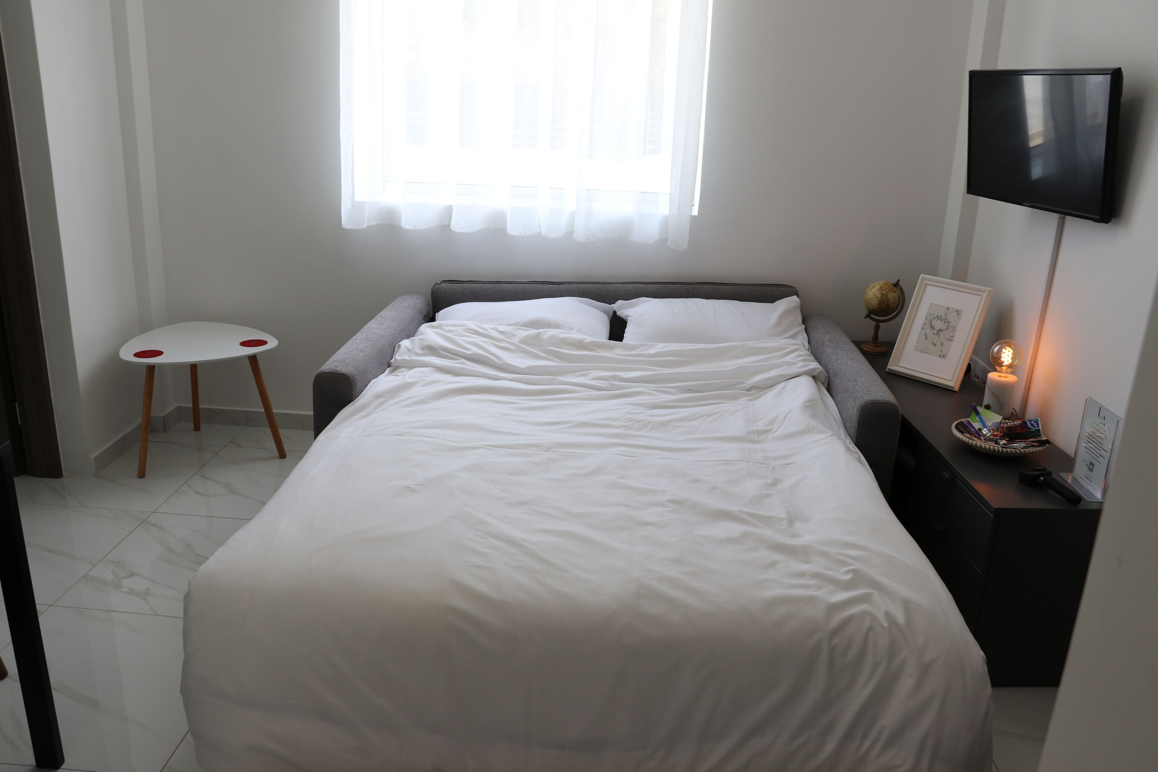 A neatly made bed with white bedding, a small white stool with red accents, a bedside table with a globe and framed picture, and a wall-mounted TV.