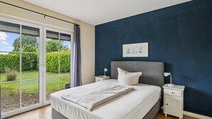 Ein Schlafzimmer mit einem großen Bett, einem Nachttisch, einem Fenster mit Blick auf Grünflächen und einer dunkelblauen Wand.