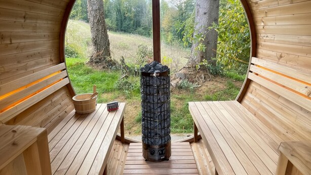 A sauna with wooden benches and a stone heater, surrounded by a wooden structure with a view of trees and grass. - Nice house with a garden and sauna. (Schinnen)