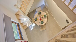 A high-ceilinged room with a wooden staircase, a round dining table with green plates, and a window with blinds. - Nice house with a garden and sauna. (Schinnen)