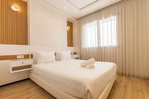 Quarto de hotel com uma cama grande, mesa de cabeceira, luminária na parede e janela com cortinas.