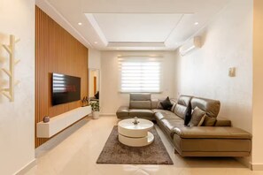 Ein modernes Wohnzimmer mit Ecksofa, Flachbildfernseher an einer Wand aus Holzpaneelen, rundem Couchtisch und großem Fenster mit Jalousien.