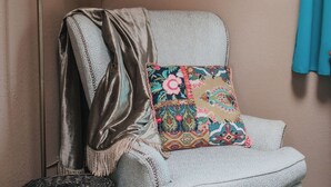 Un coin de pièce avec un fauteuil bleu clair, un coussin décoratif, une étagère fixée au mur, une lampe sur pied et un panier en osier.
