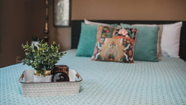 Un lit bien fait, avec une couvre-lit à motifs, un coussin décoratif et une table de chevet agrémentée d’une plante et d’un objet décoratif.