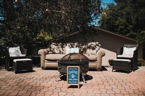 A fire pit with a sign, wicker chairs, a sofa, and a brick patio.