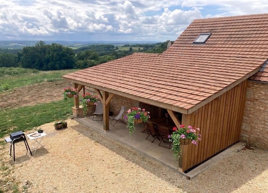 Ein überdachter Außenbereich mit Sitzgelegenheiten und Blick auf die Landschaft, ein Steinhaus und ein Ziegeldach.