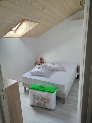 A bedroom with a sloped ceiling, a skylight, a bed with pillows, a bedside table, and a green bag on the floor.