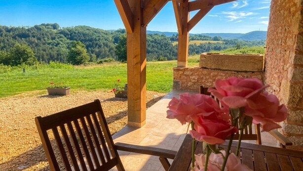 Un banc et une table en bois, placés sous une pergola adossée à un mur de pierre, offrent une vue sur un champ verdoyant et des collines.