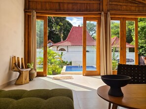 A room with a large sliding glass door, a pool, and a view of a building with a red roof. - 3-bedroom family house with pool, garden and toys (Kuta Selatan)