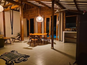 A dining area with a wooden table and chairs, a hanging light fixture, and a view of a swimming pool through glass doors. - 3-bedroom family house with pool, garden and toys (Kuta Selatan)