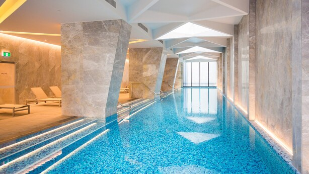 Een modern binnenzwembad met helderblauw water, marmeren wanden en een geometrisch plafondontwerp.