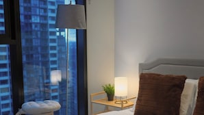 Een hotelkamer met een groot raam, een bed met opgerolde handdoeken, een nachtkastje met een lamp en uitzicht op hoge gebouwen.