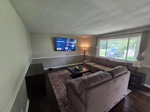 A living room with a flat-screen TV, a sofa, a coffee table, a lamp, and a window with a view of a parked car.
