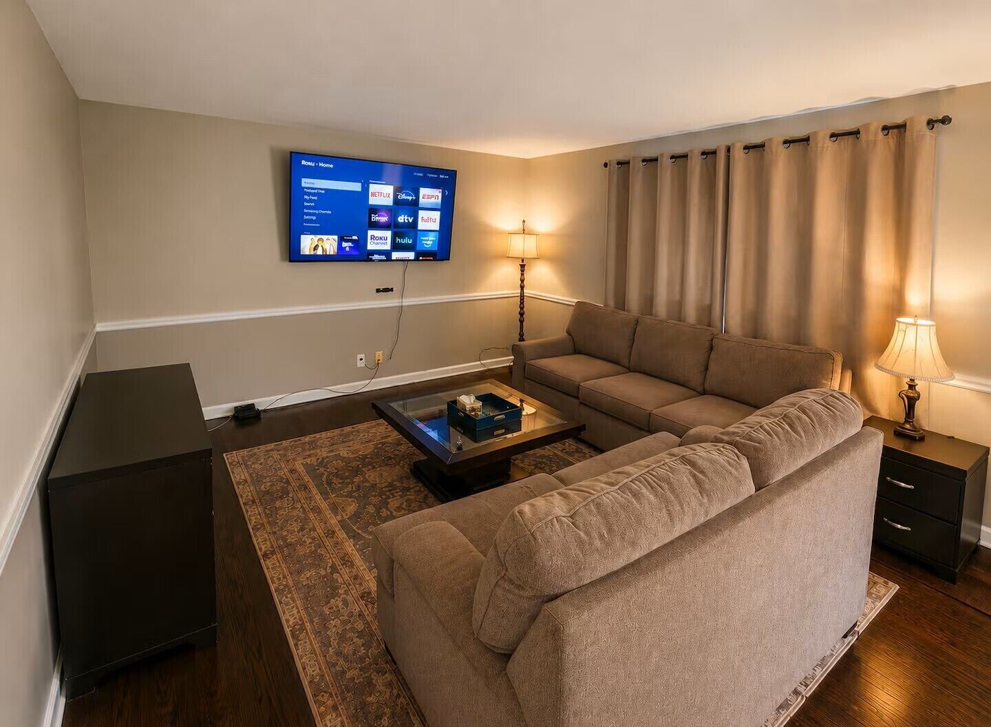 A living room with a sofa, a coffee table, a flat-screen TV mounted on the wall, a lamp, and a curtain.