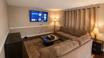 A living room with a sofa, a coffee table, a flat-screen TV mounted on the wall, a lamp, and a curtain.