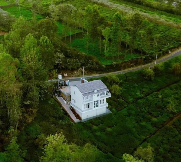 A white house with a flat roof, surrounded by lush greenery and fields.