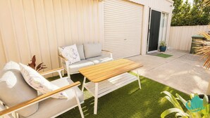 Eine Terrasse mit einem Holztisch, einem weißen Sofa, einer Schiebetür und einem kleinen Grünbereich.