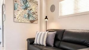 Ein schwarzes Ledersofa mit gestreiften Kissen, eine Wanduhr und eine Stehlampe in einem Raum mit einem großformatigen Kunstwerk mit Blättermotiv.