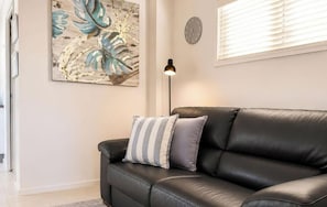 A black leather sofa with striped pillows, a wall clock, and a floor lamp in a room with a large leaf-patterned artwork. - Coastal Hideaway @ Pelican, Walk to Beach & Lake (Pelican)